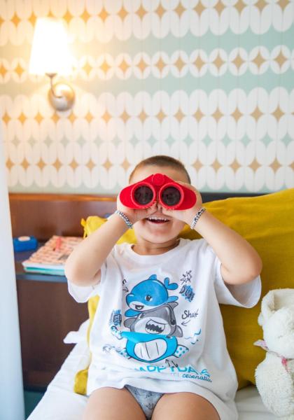 Bambino con binocolo rosso e peluche su letto.