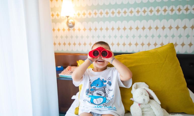 Kind mit rotem Fernglas und Plüschtier auf dem Bett.