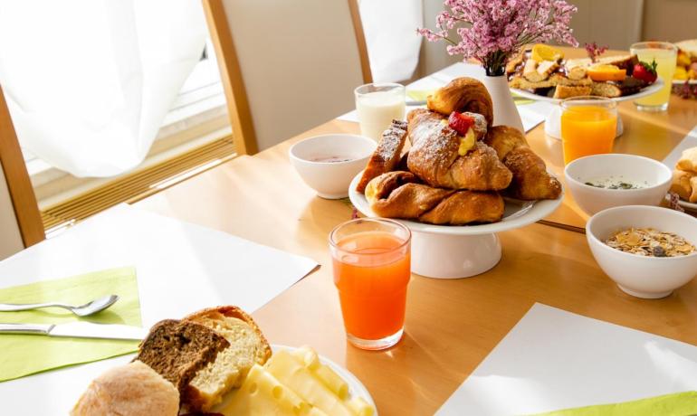 Colazione con croissant, salumi, formaggi e succhi su tavola apparecchiata.